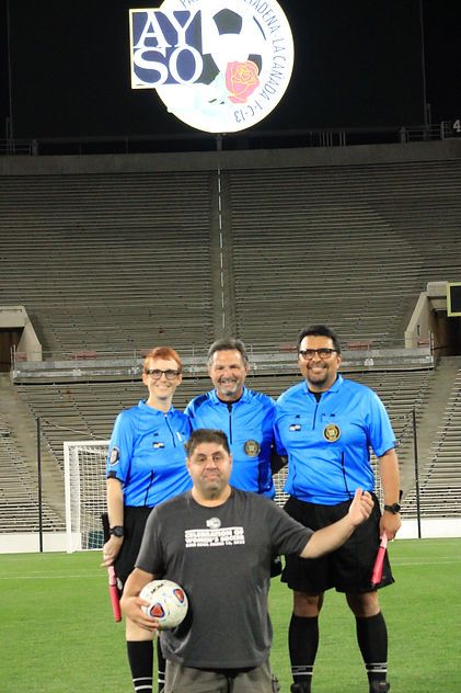 Three referees in blue uniforms posing on the Rose Bowl pitch with the AYSO Region 13 logo above them