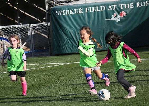 Three girls in green pinnies competing for the ball, Spieker Field banner in background