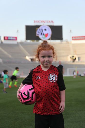 Red-haired girl in a red AYSO Region 13 jersey holding a pink soccer ball, Rose Bowl Honda jumbotron behind her